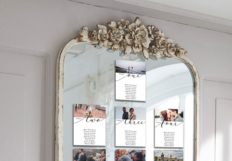 May include: A white mirror with a decorative floral frame, displaying four white cards with photos and seating arrangements for a wedding reception. The cards are labeled "Table One", "Table Two", "Table Three", and "Table Four".