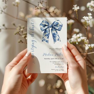 Puede incluir: Una tarjeta de invitaci&oacute;n blanca con un dise&ntilde;o floral azul y blanco. El texto en la tarjeta dice "she's tying the knot!" e incluye el nombre de la novia, la fecha, la hora y la ubicaci&oacute;n para una despedida de soltera.