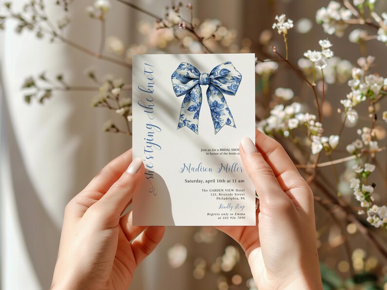 Puede incluir: Una tarjeta de invitaci&oacute;n blanca con un dise&ntilde;o floral azul y blanco. El texto en la tarjeta dice "she's tying the knot!" e incluye el nombre de la novia, la fecha, la hora y la ubicaci&oacute;n para una despedida de soltera.