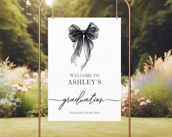 May include: A white sign with a black bow illustration and the words "WELCOME TO ASHLEY'S graduation" in script. The sign is displayed on a gold metal stand, suitable for a graduation event.