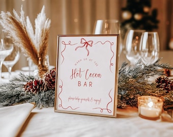 May include: A framed sign with the words "Warm Up at the Hot Cocoa Bar" in red script. The sign is surrounded by festive decorations, including pinecones, greenery, and a lit candle, creating a cozy atmosphere.