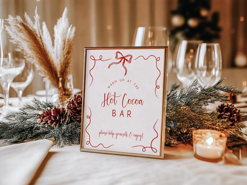 Pu&ograve; includere: Un cartello rettangolare dorato con la scritta "Warm Up at the Hot Cocoa Bar" in rosso. Il cartello &egrave; circondato da decorazioni festive, tra cui pigne, verde e una candela accesa.