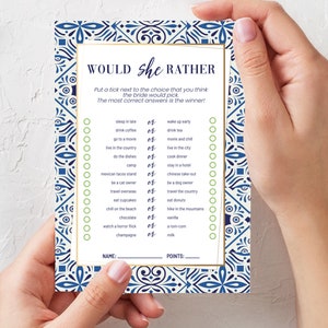 May include: A bridal shower game card with a blue and white patterned border. The card reads "Would She Rather" and includes a list of choices for the bride to select. The card is held by a person.