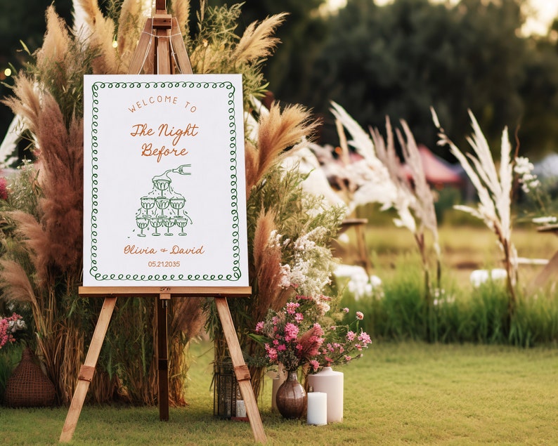 May include: A white sign with green and orange text on an easel. The sign says "Welcome to The Night Before Olivia + David 05.21.2035". The sign is decorated with a drawing of a champagne tower.