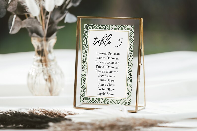 May include: A gold-framed table number card with a green and white patterned border. The card reads "table 5" and lists the names of guests assigned to that table.