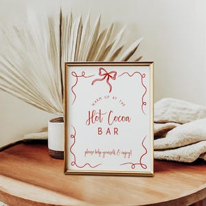 Pu&ograve; includere: Un cartello incorniciato con la scritta "Hot Cocoa Bar" in corsivo rosso. Il cartello &egrave; in una cornice dorata e presenta volute rosse decorative e un fiocco. Il cartello &egrave; su un tavolo di legno con una pianta e un panno piegato sullo sfondo.