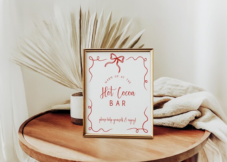 Pu&ograve; includere: Un cartello incorniciato con la scritta "Hot Cocoa Bar" in corsivo rosso. Il cartello &egrave; in una cornice dorata e presenta volute rosse decorative e un fiocco. Il cartello &egrave; su un tavolo di legno con una pianta e un panno piegato sullo sfondo.