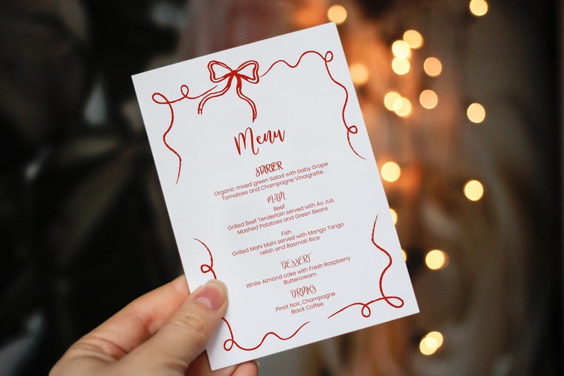 May include: A white menu with a red ribbon design. The menu lists a starter, main course, dessert, and drinks. The starter is organic mixed green salad with baby grape tomatoes and champagne vinaigrette. The main course is grilled beef tenderloin served with au jus, mashed potatoes and green beans, or grilled mahi mahi served with mango tango relish and basmati rice. The dessert is white almond cake with fresh raspberry buttercream. The drinks are pinot noir, champagne, and black coffee.