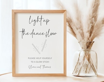 May include: A framed sign with the text "light up the dance floor" and "PLEASE HELP YOURSELF TO A GLOW STICK!" in black script. The sign is in a light wood frame and sits on a wooden table. A vase of dried flowers is to the right.