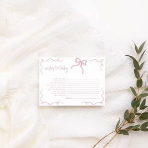 K&ouml;nnte beinhalten: Eine wei&szlig;e Karte mit einer rosa Schleife und dem Text "Wishes for baby" mit Zeilen zum Schreiben von W&uuml;nschen. Die Karte liegt auf einem wei&szlig;en Stoffhintergrund mit gr&uuml;nen Bl&auml;ttern.