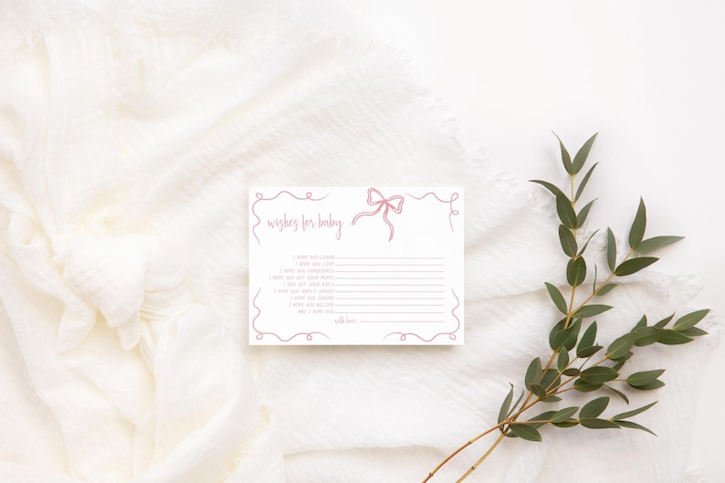 K&ouml;nnte beinhalten: Eine wei&szlig;e Karte mit einer rosa Schleife und dem Text "Wishes for baby" mit Zeilen zum Schreiben von W&uuml;nschen. Die Karte liegt auf einem wei&szlig;en Stoffhintergrund mit gr&uuml;nen Bl&auml;ttern.