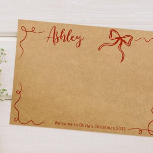 May include: A brown card with red script that says "Ashley" and "Welcome to Olivia's Christmas 2035." The card has a red bow and decorative red swirls. White flowers are in the background.