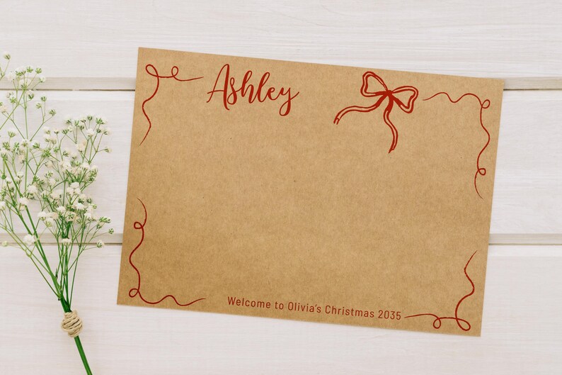 May include: A brown card with red script that says "Ashley" and "Welcome to Olivia's Christmas 2035." The card has a red bow and decorative red swirls. White flowers are in the background.
