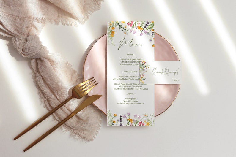 May include: A pink plate with gold trim holds a white menu card with a floral border and a place card that reads "Amanda Davenport, Table 5". The menu lists a starter, main course, and dessert. A gold fork and knife are on a beige linen napkin.