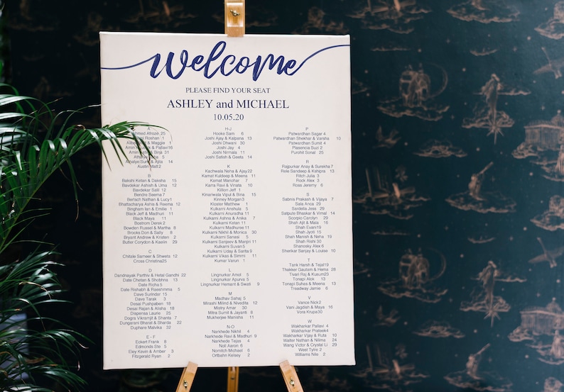 May include: A white seating chart with blue calligraphy for a wedding reception. The chart lists the names of guests and their assigned table numbers. The chart is titled "Welcome, Ashley and Michael, 10.05.20".