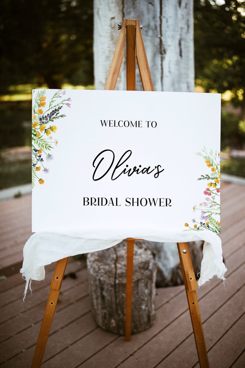 May include: A white sign on a wooden easel with the text "WELCOME TO Olivia's BRIDAL SHOWER." Floral watercolor designs in yellow, purple, and white adorn the corners. A white fabric drapes across the bottom of the sign.