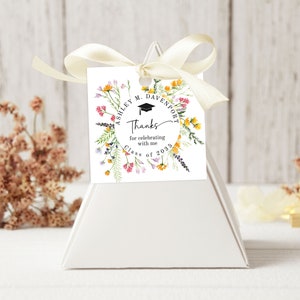 May include: White favor box with a white tag tied with a cream ribbon. The tag has a floral wreath with a graduation cap and the text "Ashley M. Davenport Thanks for celebrating with me Class of 2023".