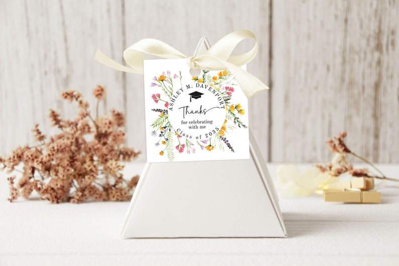 May include: White favor box with a white tag tied with a cream ribbon. The tag has a floral wreath with a graduation cap and the text "Ashley M. Davenport Thanks for celebrating with me Class of 2023".