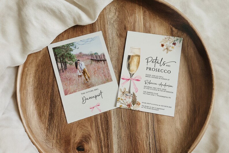 May include: Two cards on a wooden surface. The card on the left has a photo of a couple kissing in a field. The text on the card reads "The future Mrs. Davenport". The card on the right has a watercolor illustration of a glass of champagne with pink ribbon and flowers. The text on the card reads "Petals and Prosecco. Please join us for a bridal shower in honor of Rebecca Anderson. Saturday, May 23rd, 2020 at five o'clock in the afternoon. The Garden Club. 123 Park Lane | Philadelphia, PA."