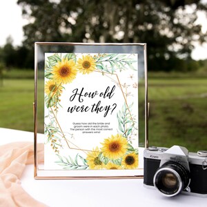May include: A white sign with a gold geometric frame and sunflower illustrations. The sign reads "How old were they?" and includes instructions for a wedding game.