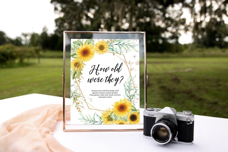 May include: A white sign with a gold geometric frame and sunflower illustrations. The sign reads "How old were they?" and includes instructions for a wedding game.