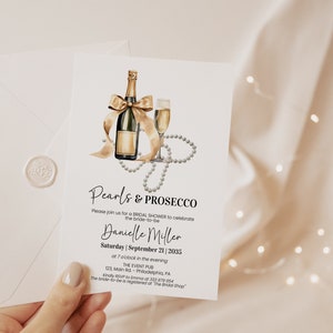 May include: A white invitation card with gold lettering for a bridal shower. The invitation features a watercolor illustration of a champagne bottle with a bow, a champagne flute, and a pearl necklace. The text on the card reads "Pearls & Prosecco" and includes the bride-to-be's name, the date, and the time of the event.