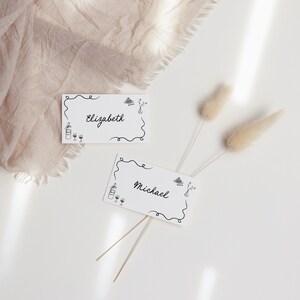 May include: Two white place cards with black calligraphy and simple illustrations. The cards read "Elizabeth" and "Michael".