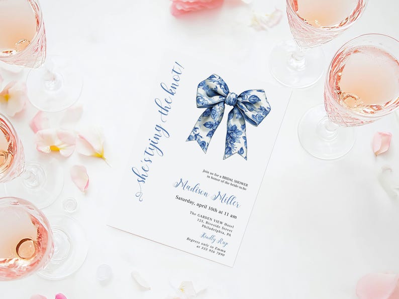Puede incluir: Un lazo floral azul y blanco sobre un fondo blanco. El texto "she's tying the knot!" est&aacute; escrito en cursiva azul en el lado izquierdo de la imagen. El texto "Madison Miller" est&aacute; escrito en cursiva azul debajo del lazo. El texto "Saturday, April 10th at 11 am" est&aacute; escrito en azul debajo del nombre. El texto "The Garden View Hotel" est&aacute; escrito en azul debajo de la fecha. El texto "133 Riverside Street, Philadelphia, PA" est&aacute; escrito en azul debajo del nombre del hotel. El texto "Kindly RSVP" est&aacute; escrito en azul debajo de la direcci&oacute;n. El texto "Regrets only to Emma at 215-555-1212" est&aacute; escrito en azul debajo de la RSVP.