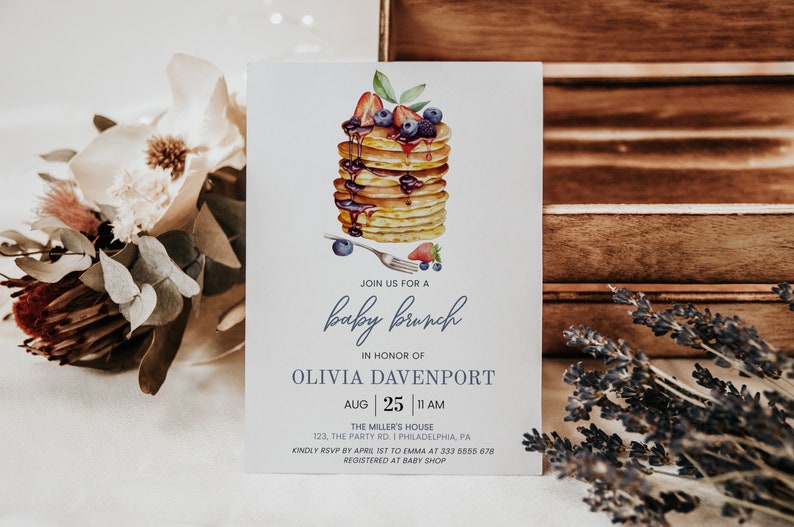 May include: A baby shower invitation with a watercolor illustration of a stack of pancakes topped with blueberries, strawberries, and syrup. The text reads "Join us for a baby brunch in honor of Olivia Davenport, Aug 25, 11 AM, The Miller's House, 123, The Party Rd. | Philadelphia, PA, Kindly RSVP by April 1st to Emma at 333 5655 678, Registered at Baby Shop."