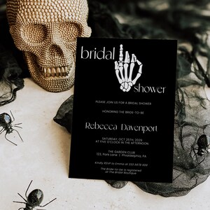 K&ouml;nnte beinhalten: Eine schwarz-wei&szlig;e Einladung zur Bridal Shower mit einer Skeletthand, die einen Ring h&auml;lt. Die Einladung lautet "bridal shower" und enth&auml;lt den Namen der Braut, das Datum, die Uhrzeit und den Ort der Veranstaltung. Au&szlig;erdem enth&auml;lt sie die RSVP-Informationen.