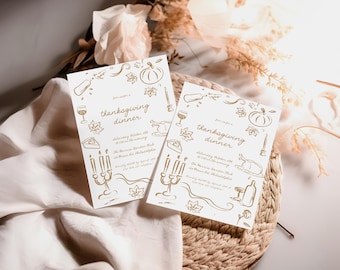 May include: Two white cards with gold lettering and illustrations of Thanksgiving dinner elements, such as a turkey, pumpkin, wine glass, and candles. The text on the cards reads "Thanksgiving Dinner" and includes the date and time of the event. The cards are on a woven straw placemat.