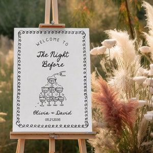 May include: White sign with black text and a drawing of champagne glasses. The text reads "Welcome to The Night Before Olivia + David 05.21.2035".
