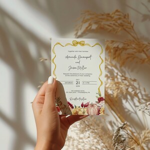 May include: A wedding invitation with a gold ribbon and bow, featuring a floral design with pumpkins, leaves, and flowers in shades of red, orange, and yellow. The text reads "Amanda Davenport and Jason Miller" with the date and time of the wedding: "Saturday, October 21, 2019 at 5 PM". The location is "The Garden Hotel, 123 Pine Lane, Philadelphia, PA".