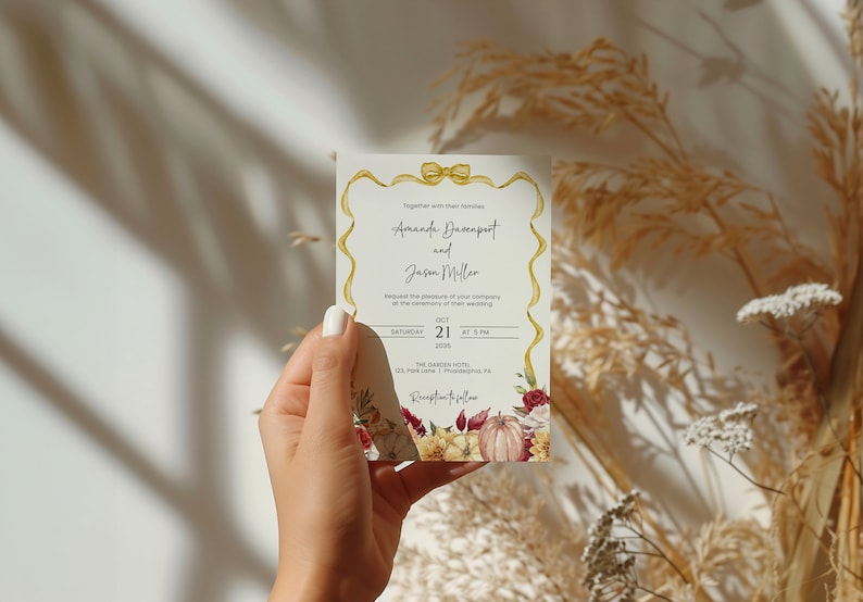 May include: A wedding invitation with a gold ribbon and bow, featuring a floral design with pumpkins, leaves, and flowers in shades of red, orange, and yellow. The text reads "Amanda Davenport and Jason Miller" with the date and time of the wedding: "Saturday, October 21, 2019 at 5 PM". The location is "The Garden Hotel, 123 Pine Lane, Philadelphia, PA".