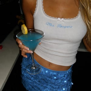 Può includere: Una canotta bianca a coste con bordo in pizzo, un piccolo fiocco e il testo ricamato in blu "Mrs. Recupero" sul petto, abbinata a una minigonna di paillettes blu brillante. Un bicchiere da martini con liquido blu e una fetta di limone.