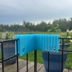 May include: A bright blue, L-shaped plastic corner connector piece with vertical ribbed texture and two circular holes on each side, designed to fit over the top corner of a black wire grid panel structure for stability and alignment.
