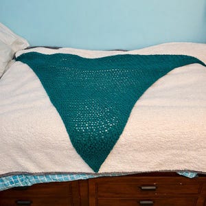 May include: A triangular teal crochet shawl featuring an openwork stitch pattern. The accessory is crafted with a consistent yarn texture and displays a wide top edge that tapers down to a single point at the bottom, showcasing a handmade design.