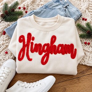 May include: A cream-colored crewneck sweatshirt featuring the word Hingham embroidered in a textured, bright red script font across the chest. The garment has long sleeves, a ribbed neckline, and a soft, fleece-like interior lining.