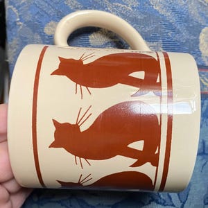 May include: A cream-colored ceramic mug featuring a repeating pattern of rust-colored cat silhouettes. The design is framed by vertical rust-colored lines on both sides. The text 1992 is visible near the top handle attachment point.