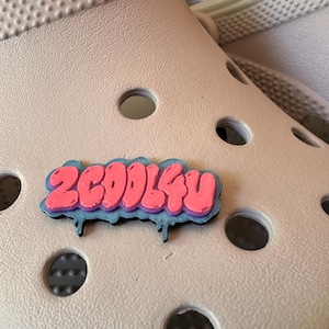 May include: A shoe charm featuring the text 2COOL4U in a pink graffiti-style font with a light blue outline and black drip accents. The charm is attached to a beige foam clog with circular ventilation holes, showcasing a textured surface.