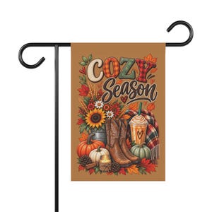 May include: A rectangular garden flag featuring the text "COZY SEASON" in stylized lettering. The design includes a sunflower bouquet, two brown cowboy boots, pumpkins, a candle, a plaid blanket, and a coffee cup on a tan background.