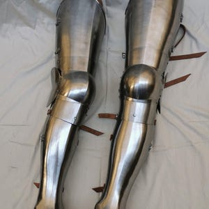 May include: A pair of polished steel leg armor pieces, known as greaves and cuisses, featuring articulated knee cops, metal rivets, and brown leather straps for fastening. The metallic surface reflects light, showing the curved, segmented construction.