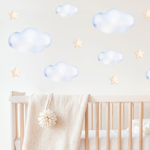 May include: A set of wall decals featuring light blue watercolor-style clouds and pale yellow stars. The decals are arranged on a wall above a light wood crib draped with a cream-colored knit blanket. The text PRINTBEE is visible.