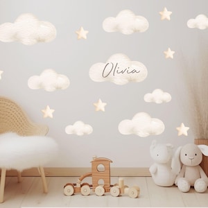 May include: A set of wall decals featuring beige clouds and stars, with one cloud displaying the name Olivia in cursive script. The decals are arranged on a wall above a wooden toy train and plush toys, with the text PRINTBEE visible.