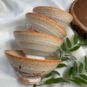 May include: A stack of four ceramic bowls with a speckled cream and orange glaze. Each bowl features a wide, flared rim and a tapered base, with a hand-painted brown botanical design visible on the exterior of the bottom bowl.