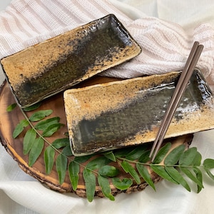May include: Two rectangular ceramic plates with a speckled tan and dark brown glazed finish. The plates are arranged on a wooden board with green leaves, and a pair of brown chopsticks rests on the lower plate, creating a set for dining.