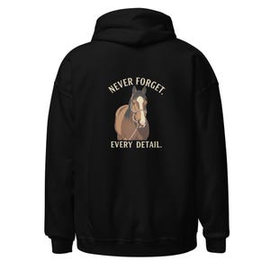 Puede incluir: Una sudadera con capucha negra que presenta un gráfico de un caballo marrón con una mancha blanca en la cara, usando un cabestro. El texto "NEVER FORGET." está impreso en color beige claro sobre el caballo y "EVERY DETAIL." debajo.