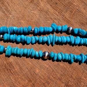 May include: A multi-strand necklace featuring small, irregular, bright turquoise blue stone chips interspersed with smooth, round, polished silver-toned metal beads. The necklace is arranged in three horizontal rows across the frame.