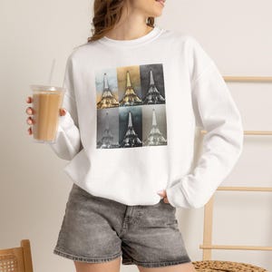 May include: A white long-sleeve sweatshirt featuring a graphic print of the Eiffel Tower in six distinct panels. Each panel displays the tower in a different color tone, ranging from warm golden hues to black and white and cool gray variations.