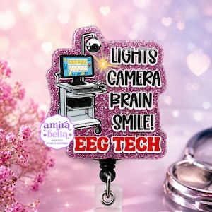 May include: A pink glitter badge reel featuring an illustration of an EEG machine with a monitor displaying brain waves. The text reads LIGHTS CAMERA BRAIN SMILE! EEG TECH in black and red lettering. A small logo for amira bella is visible.